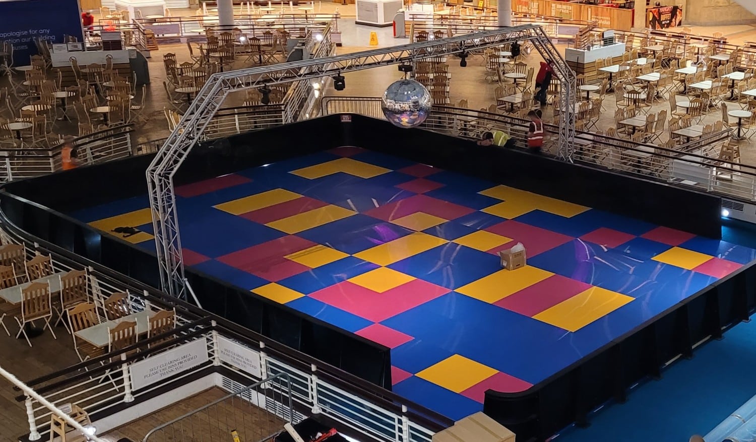ComicRelief A colorful roller rink with a disco ball, surrounded by empty tables and chairs in a food court.