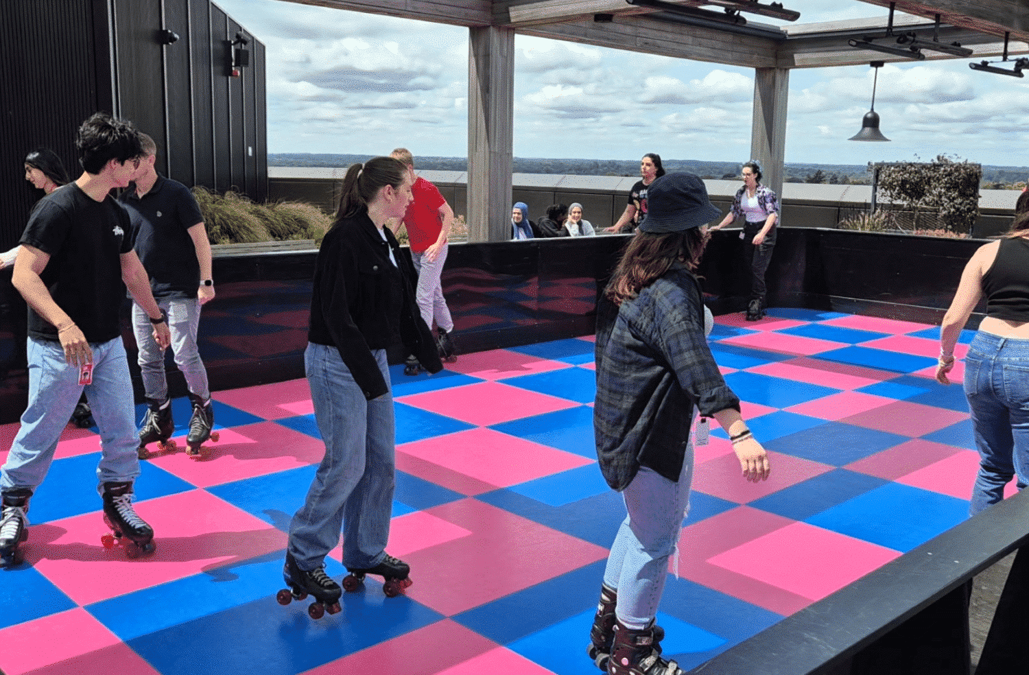 ComicReliefRoller People roller skating on a rooftop rink with a pink and blue checkered floor under a cloudy sky.
