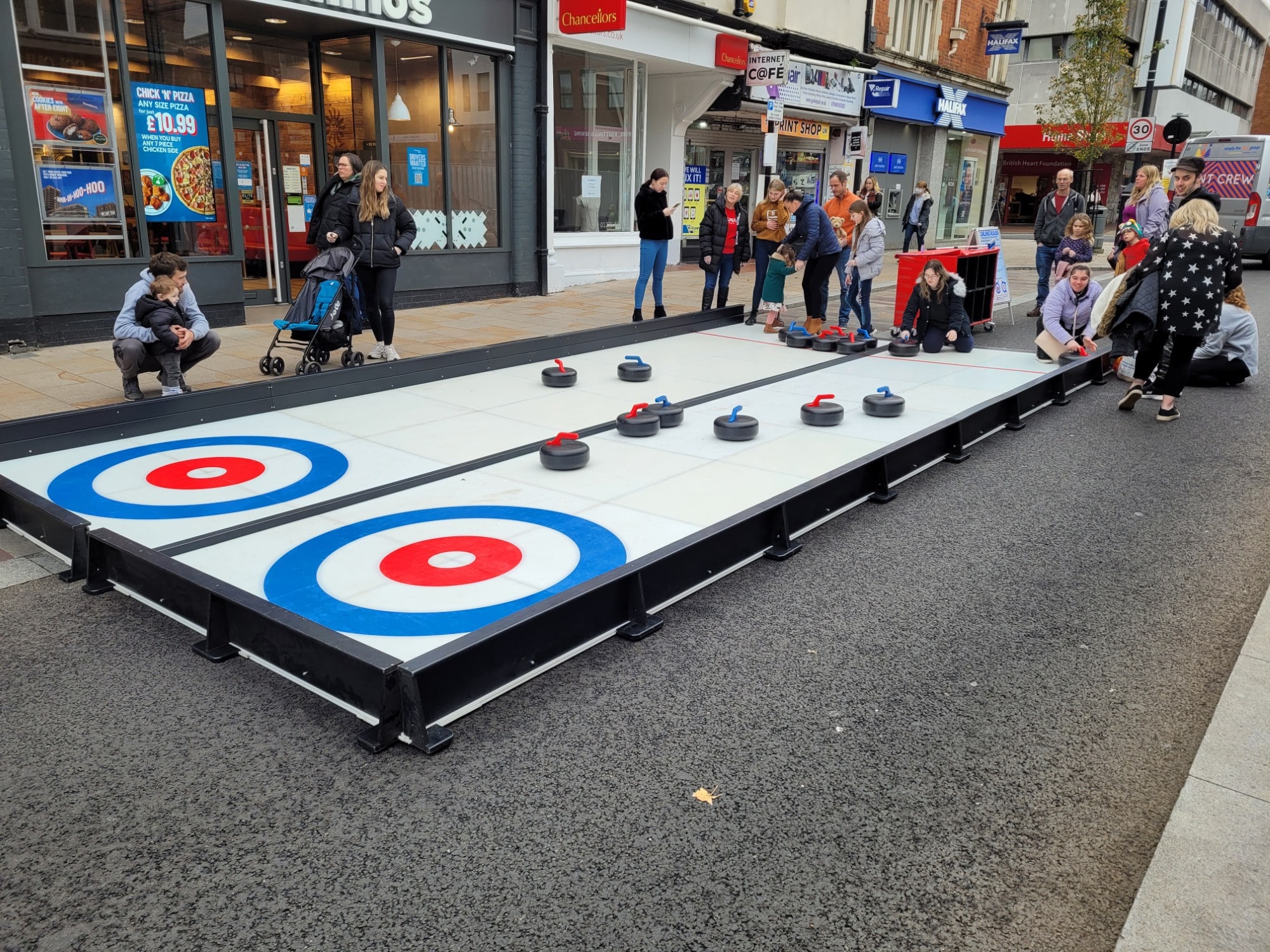 Curling People play street curling on a portable rink in a busy town center, with onlookers watching.