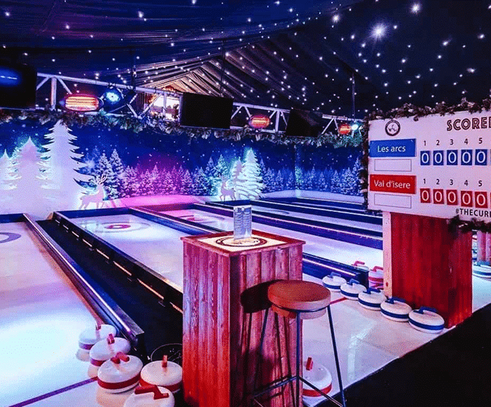 Indoor curling lanes with a festive winter theme, scoreboard, and decorated trees.