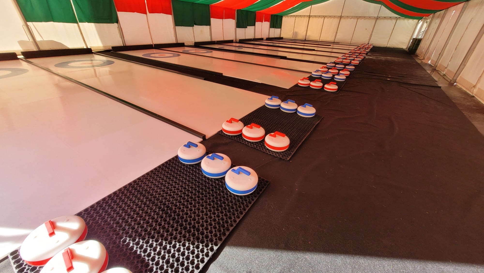 CurlingLanes Several indoor curling lanes with red and blue curling stones on mats under a striped tent ceiling.