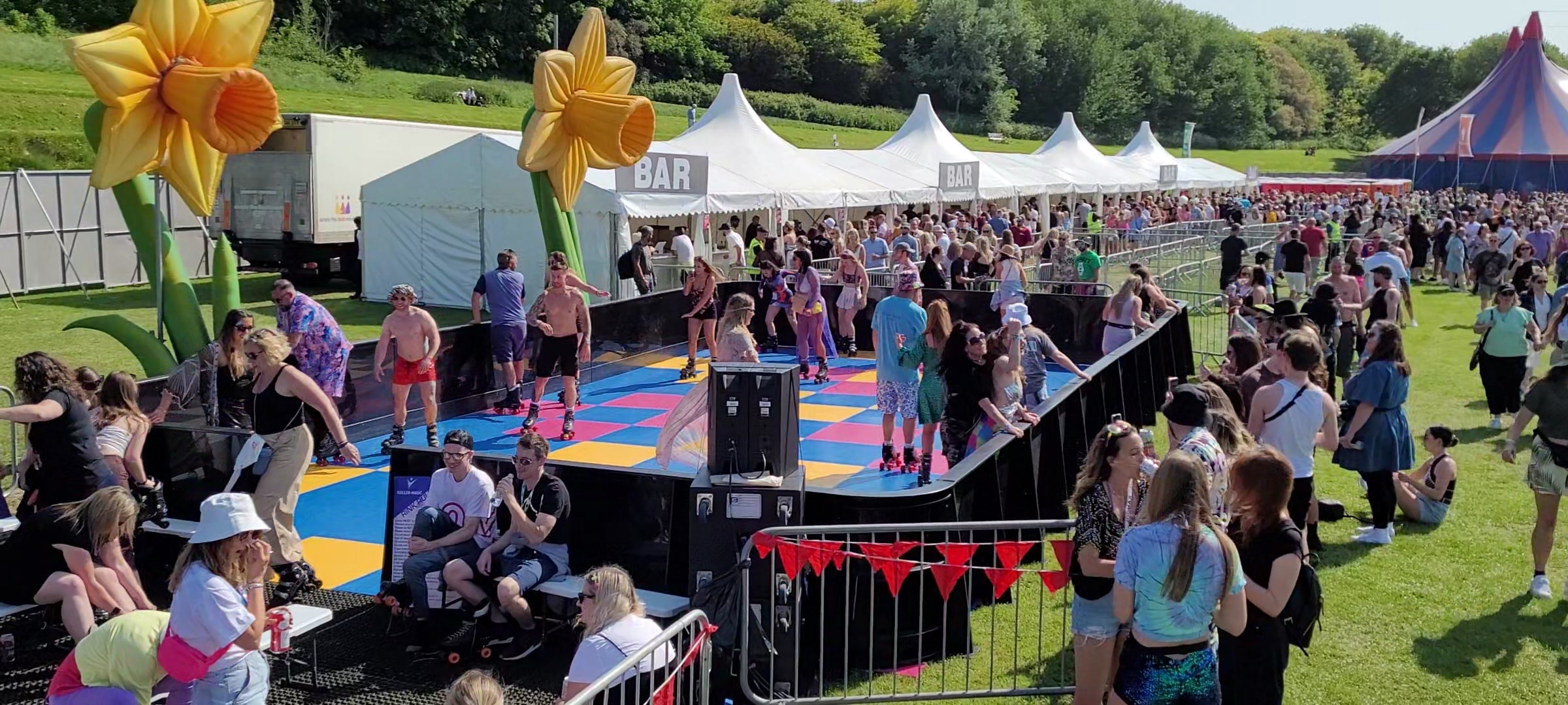 People skating on a colorful outdoor roller rink at a festival with crowds and tents in the background.