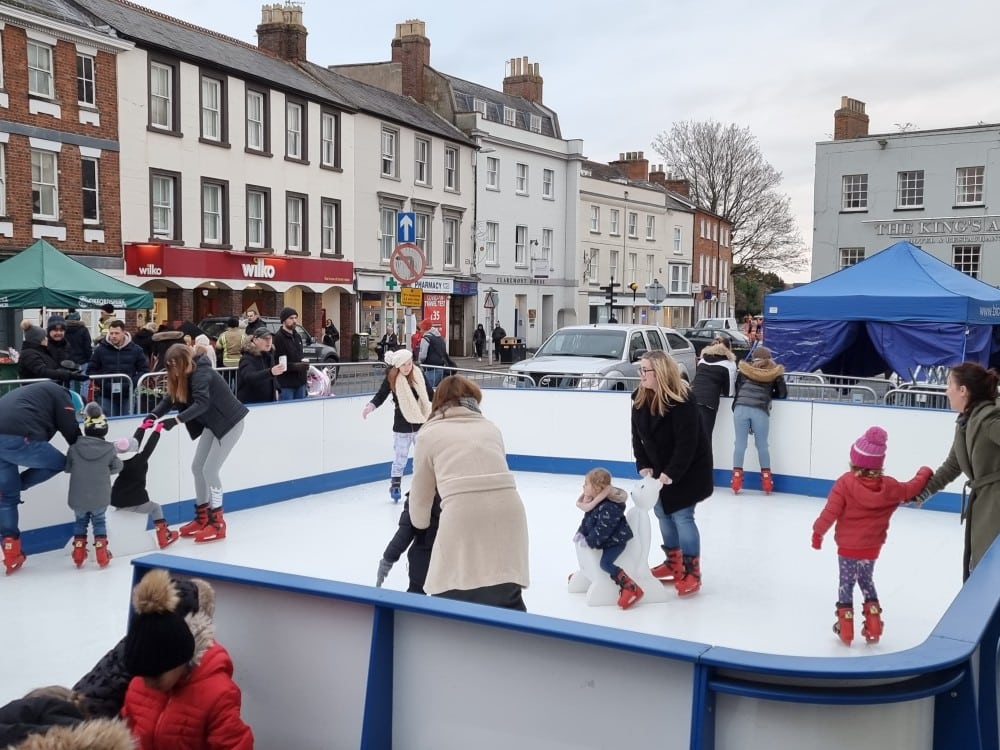 Children and adults ice skating at a town centre outdoor rink with buildings in the background.
