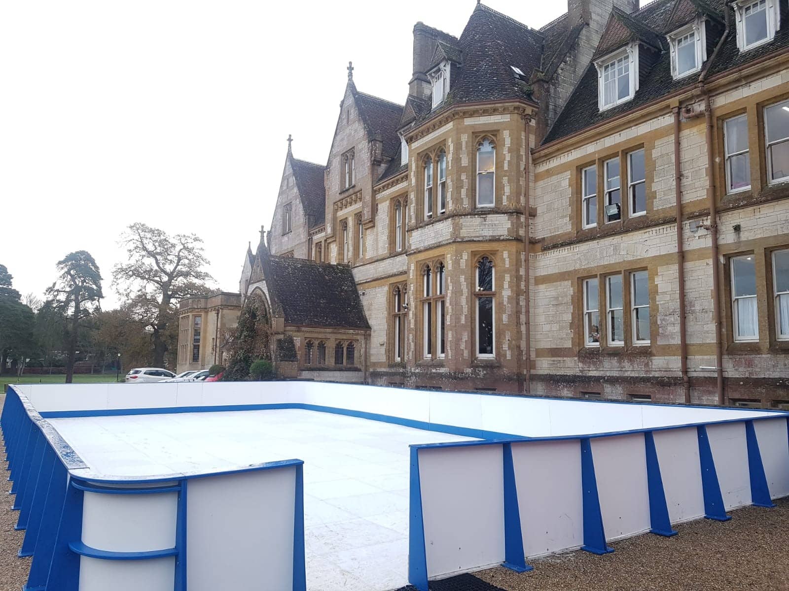 Small outdoor ice rink with blue boards in front of a large historic building.