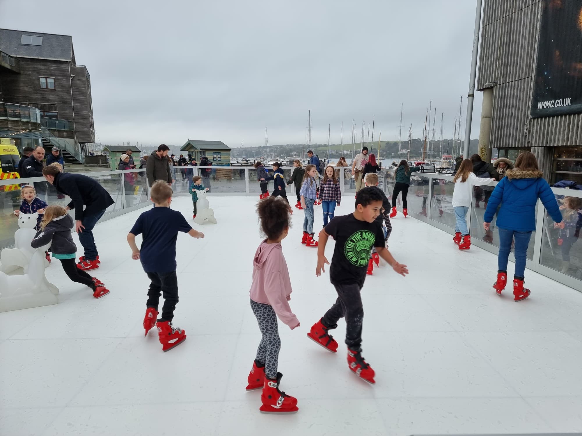 Children and adults ice skating outdoors on a white rink with red skates.