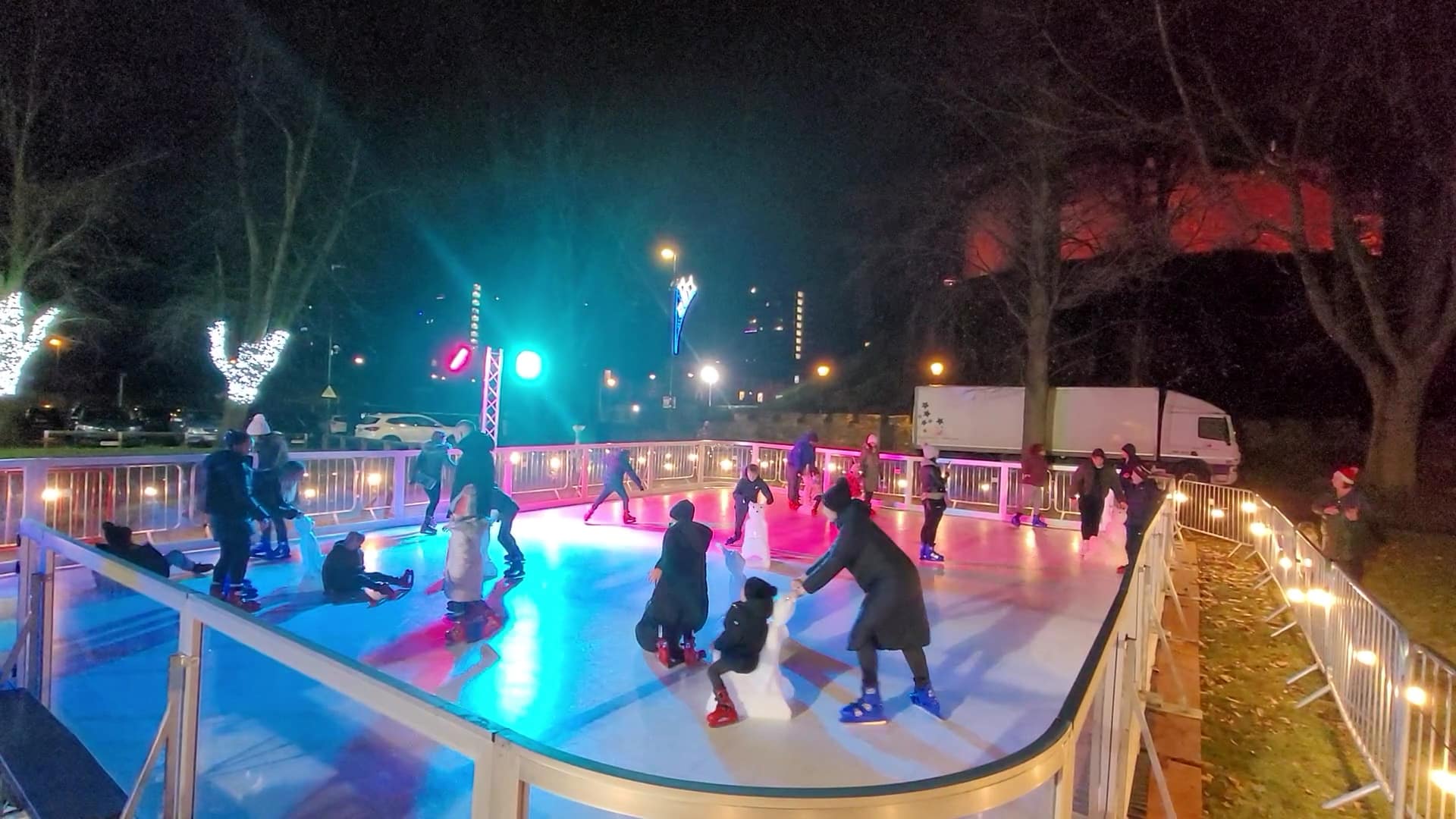 People ice skating at an outdoor rink at night with festive lights and trees in the background.