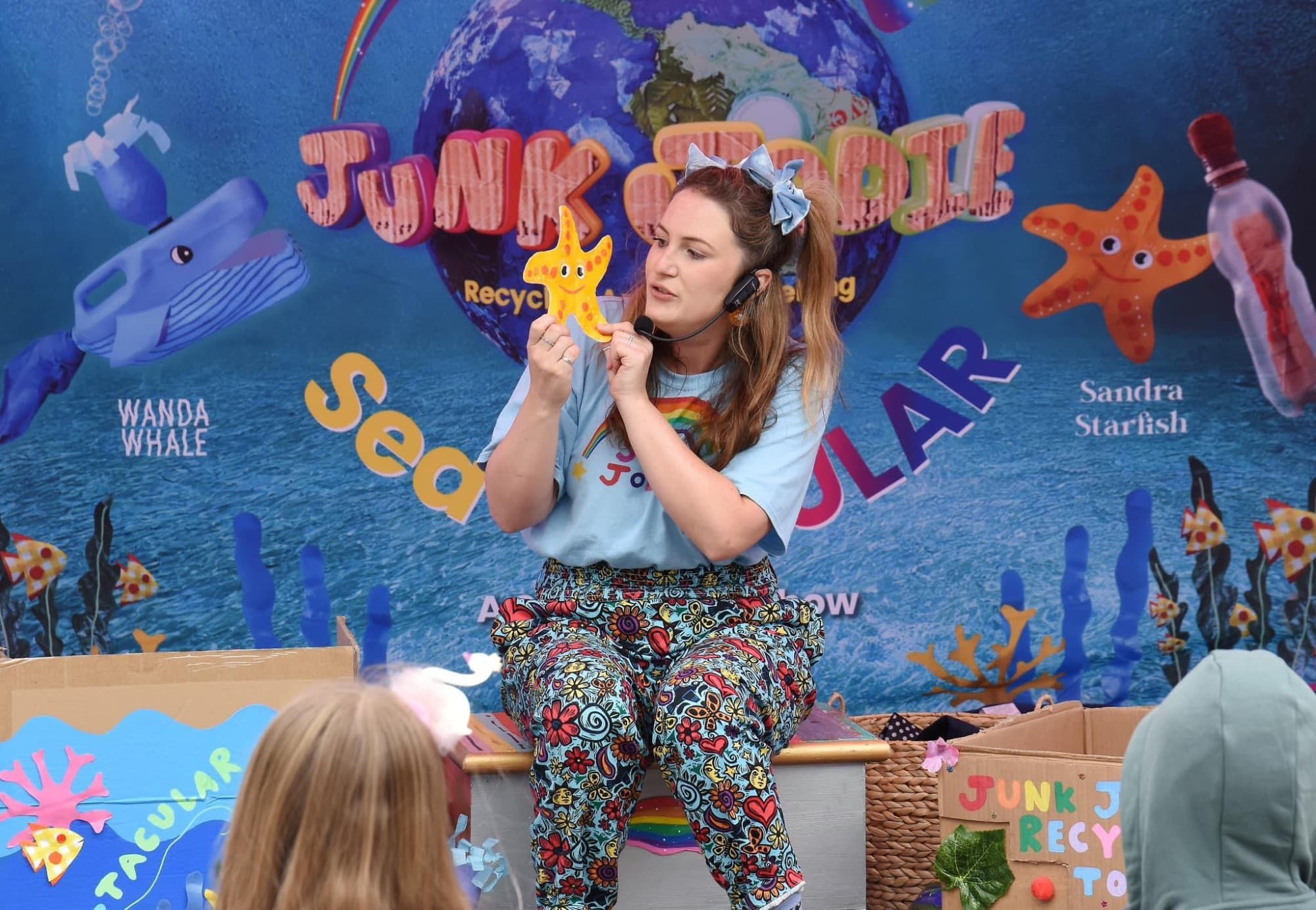 JunkJodie Woman with a headset holds a starfish craft on stage, with colorful ocean-themed backdrop and boxes.