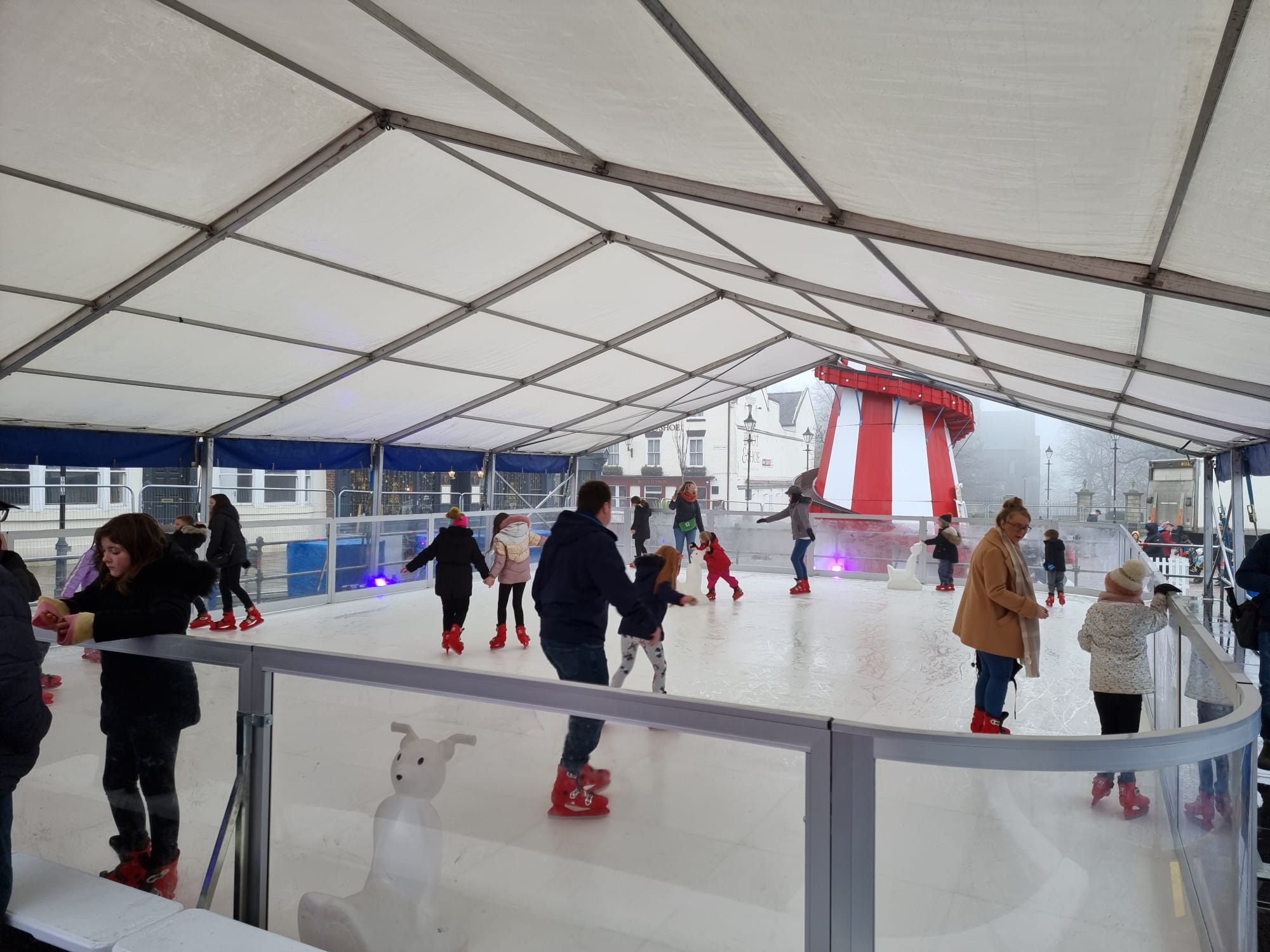 People ice skating on an outdoor rink under a large white marquee tent.