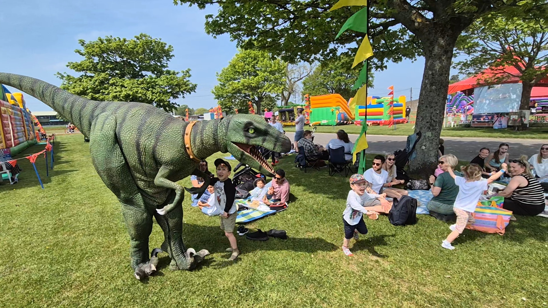 MeganFamilyFunFest-Dinosaur Children play near a person in a dinosaur costume at an outdoor family festival with bouncy castles.