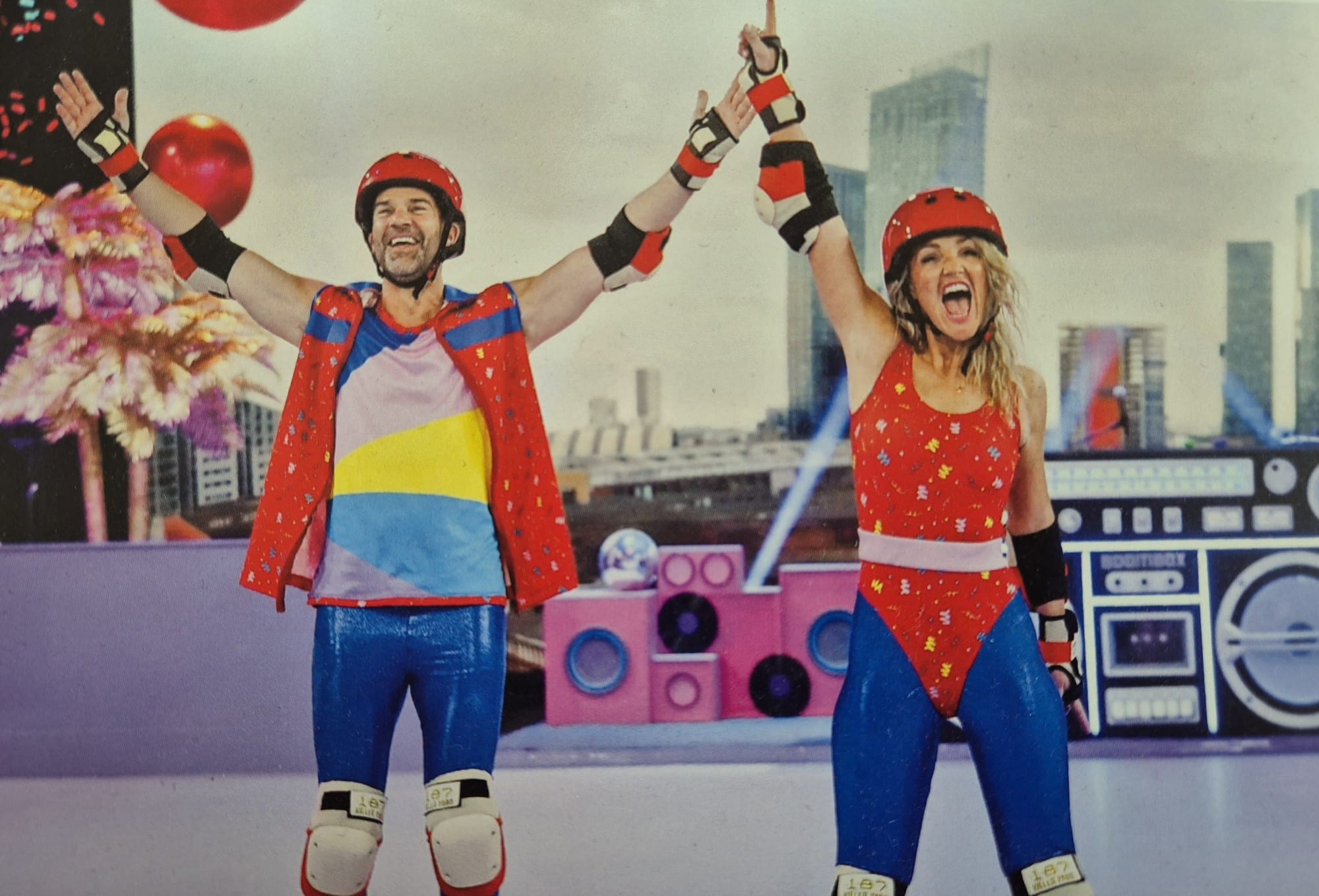 RollerRink - ComicRelief Two people in colorful roller disco outfits celebrate on a rink with their arms raised.