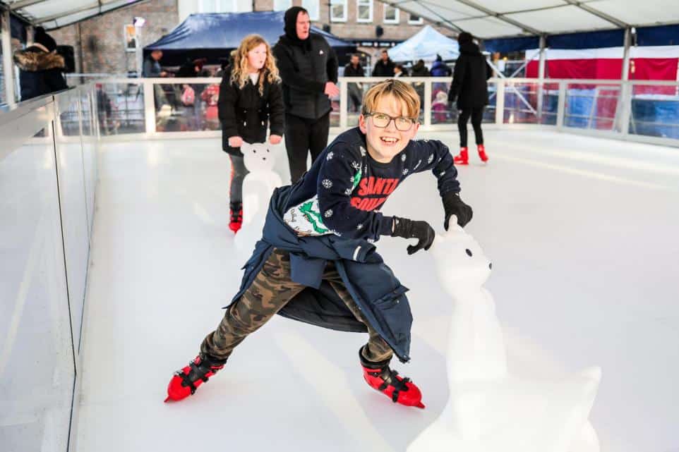 Boy in glasses ice skating with a polar bear skating aid at an indoor rink.