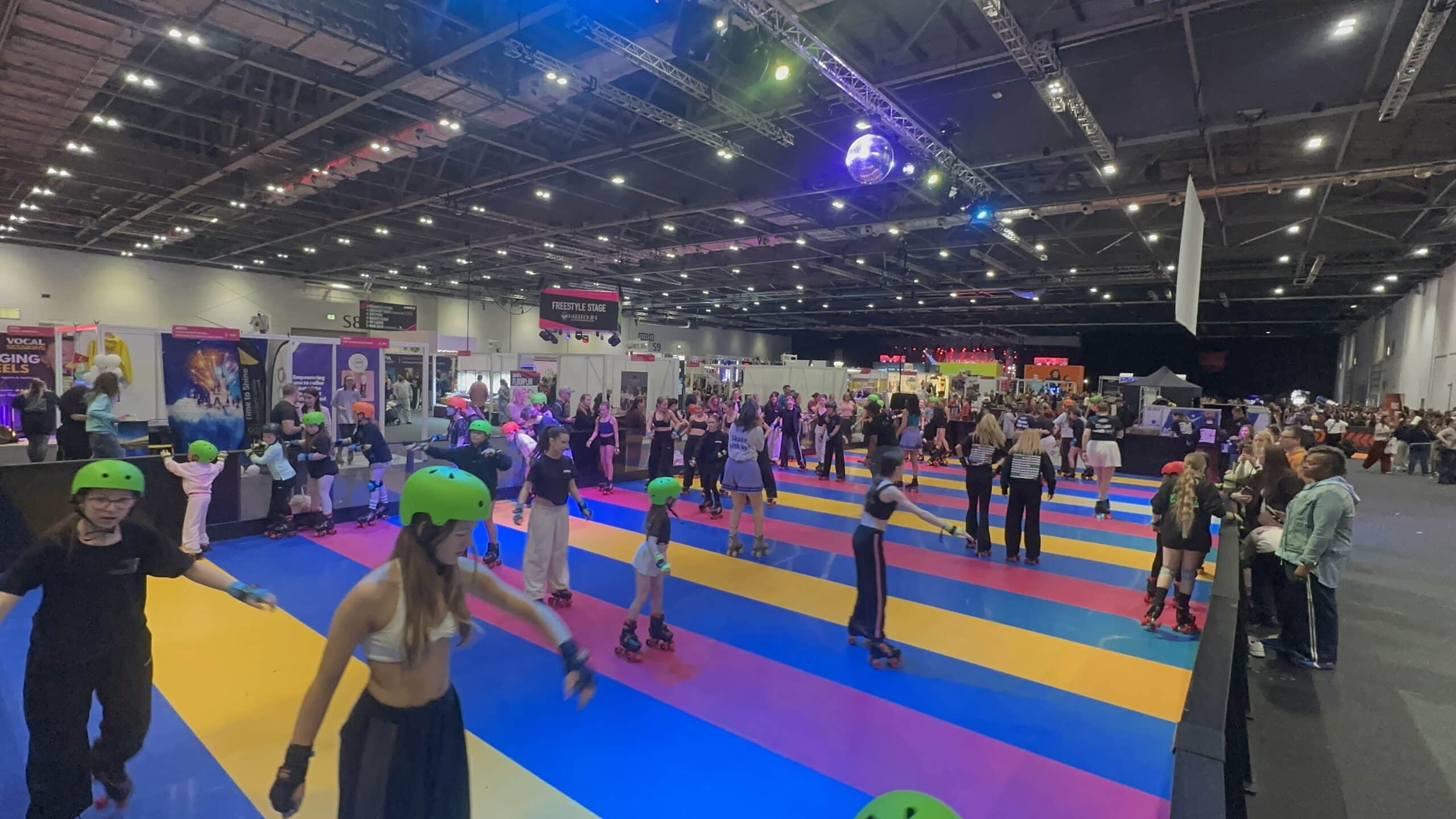 People roller skating in a colorful indoor rink with bright lights, palm trees, and large pink balloons.