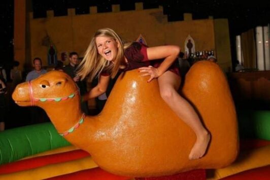 Woman riding a mechanical camel at an indoor event, smiling and having fun.