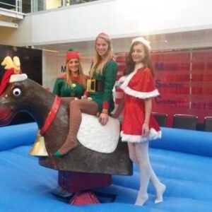Three women in Christmas costumes stand and sit on a mechanical reindeer in an indoor setting.