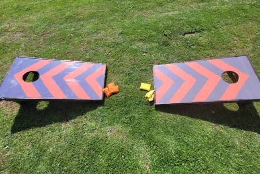 Two cornhole boards with orange and yellow bean bags on grass.