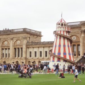 People gather on a lawn near a large historic building and a red and white helter skelter fairground ride.