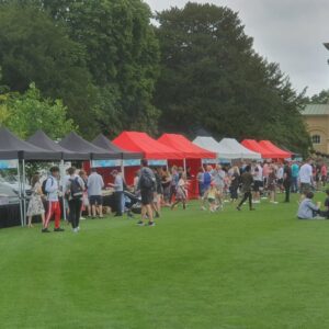 People at an outdoor event with black, red, and white gazebos on a grassy lawn.
