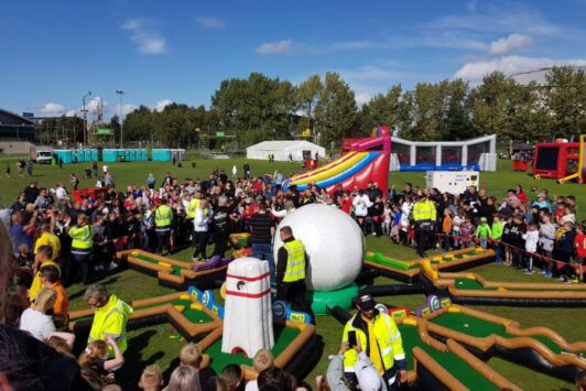 Crowd gathers around an inflatable crazy golf course at an outdoor event on a sunny day.