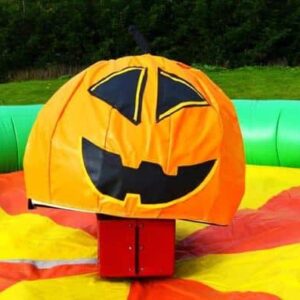 Large inflatable pumpkin ride sits on colorful mat, outdoors with green grass and trees in background.