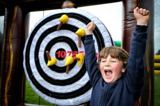 Smiling child celebrating in front of an inflatable axe-throwing target with foam axes stuck to it.
