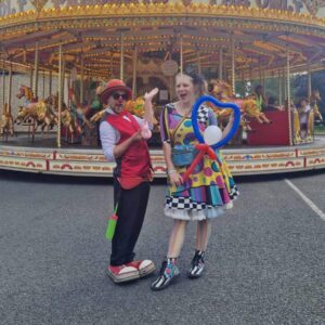 Two entertainers in colorful costumes stand in front of a carousel at a fair.