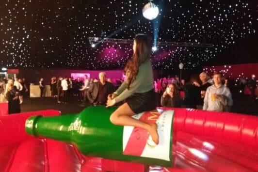 A woman rides a mechanical bottle at a party with people and lights in the background.