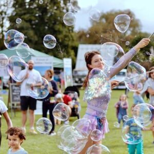 Woman in a sparkly outfit making large bubbles outdoors, with children and adults watching.