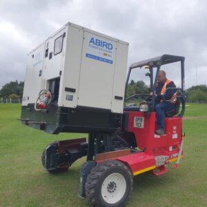 Man operating a red forklift carrying a large ABIRD generator on a grassy field.