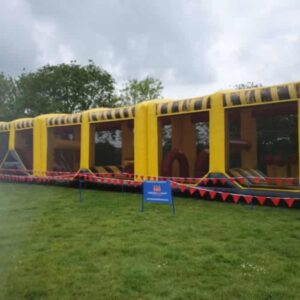 Large yellow inflatable obstacle course outdoors on grass, surrounded by red bunting and a blue sign.