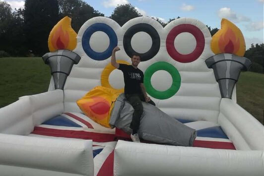 Man on inflatable Olympic torch ride with Olympic rings and torches in the background outdoors.