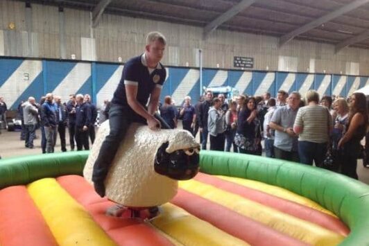 A man rides a mechanical sheep at an indoor event with people watching.