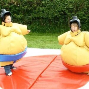 Two children in sumo wrestling suits stand on a mat outdoors, smiling at each other.