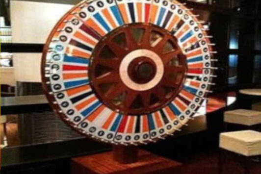 Large colorful spinning casino wheel with numbers, set up indoors near bar stools and tables.