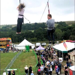 Two people riding a zip wire over a festival crowd with tents and green fencing below.